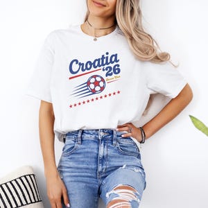 A woman wearing a white t-shirt with a graphic of a soccer ball and the text "Croatia 26" stands next to a potted plant.