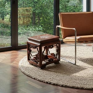 Puede incluir: Escena de un salón moderno con un sillón de cuero marrón con estructura cromada, una pequeña mesa auxiliar de madera y una alfombra redonda de yute. Una estantería empotrada y una gran ventana con vistas a árboles completan el interior.
