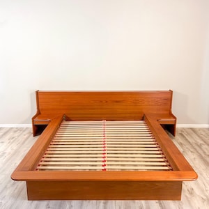 May include: A teak wood bed frame featuring an integrated headboard that extends into two attached side tables. The bed base includes a slatted support system with light-colored wooden slats and red plastic connectors for mattress placement.