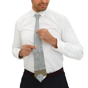 May include: A light grey tie with a detailed ruler design. The tie has a beige-coloured bottom section and is worn with a white dress shirt and black trousers. The tie is being adjusted by the wearer.
