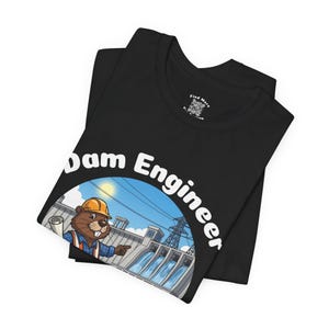 Puede incluir: Camiseta negra con un castor de dibujos animados con casco que señala una ilustración de una presa. El texto "Dam Engineer" está arqueado sobre la imagen. La camiseta está doblada, mostrando el cuello y un pequeño logotipo.