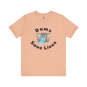 May include: Peach-colored t-shirt featuring the text "Dams Save Lives" in a black, rounded font. The graphic depicts a cartoon dam with water flowing through it. The shirt is made of a soft material.