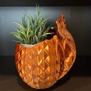 May include: A decorative, copper-colored planter shaped like a dragon egg, with a detailed scale pattern. A dragon figure is attached to the side, with its mouth open. The planter holds green artificial plants.