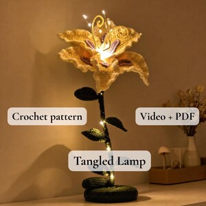 May include: A crocheted flower lamp with a yellow and white flower head, green stem, and base. The stem is adorned with small green leaves and fairy lights. The text "Tangled Lamp", "Crochet pattern", and "Video + PDF" are visible.