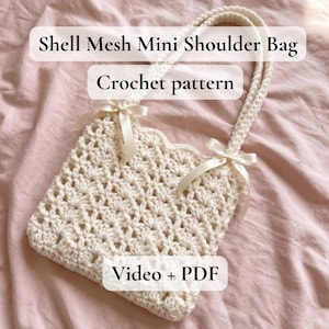 May include: A cream-colored, crocheted shell mesh mini shoulder bag with a scalloped edge. The bag features a woven handle and is adorned with cream-colored bows. Text on the image reads "Shell Mesh Mini Shoulder Bag", "Crochet pattern", and "Video + PDF".
