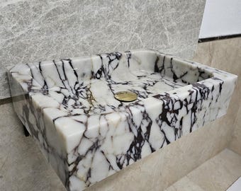 May include: A white marble sink with purple veins. The sink is rectangular and has a slightly rounded edge. It is mounted on a wall and has a small drain hole in the center.