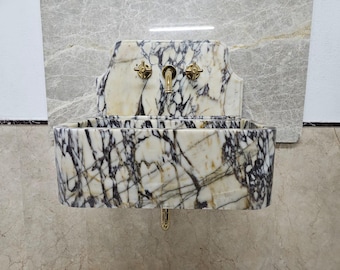 May include: A marble wall-mounted sink with brass fixtures. The sink basin is rectangular with a white and black veined pattern. The faucet and two handles are brass. The sink is mounted on a light gray marble wall.