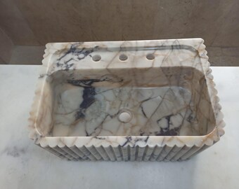 May include: Rectangular marble sink with a scalloped edge. The sink has a light beige background with dark gray and black veining. It features three faucet holes and a central drain.