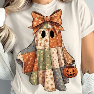 May include: A cream t-shirt with a patchwork ghost design. The ghost has a large orange bow, black eyes, and a patchwork body with floral patterns in orange, green, and blue. It holds an orange jack-o'-lantern bucket, ideal for Halloween.