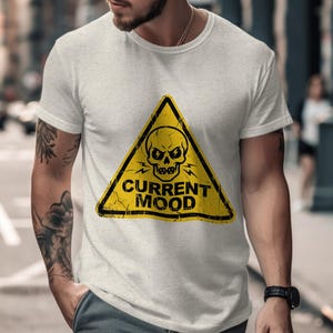 以下が含まれることがあります： 黄色の三角形にスカルのグラフィックと黒い文字「CURRENT MOOD」がプリントされた、薄いグレーのTシャツ。Tシャツは、腕にタトゥーのある人が着ています。