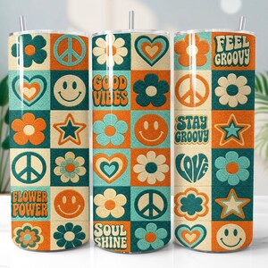 May include: Three cylindrical tumblers with straws, each featuring a grid pattern of squares containing orange, teal, and cream-colored icons including flowers, peace signs, hearts, stars, and smiley faces, with text reading "Feel Groovy," "Good Vibes," "Stay Groovy," "Love," "Flower Power," and "Soul Shine."