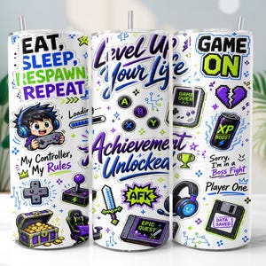 May include: A white cylindrical tumbler featuring a vibrant gaming-themed design with purple, green, and black graphics. The print includes text like EAT, SLEEP, RESPAWN, REPEAT, LEVEL UP YOUR LIFE, and GAME ON, alongside icons of controllers, a treasure chest, a sword, and a floppy disk.