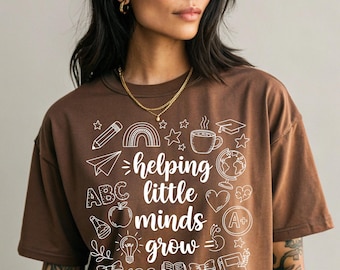 Może przedstawiać: Brązowa koszulka z białym napisem „helping little minds grow”, otoczona szkolnymi ilustracjami: ołówkiem, tęczą, jabłkiem i książkami. Koszulka ma luźny krój.