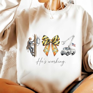 Op de afbeelding: Witte sweatshirt met een afbeelding van een elektricien op een elektriciteitspaal, een strik en een utility truck met een Amerikaanse vlag. De tekst "He's working" staat eronder.
