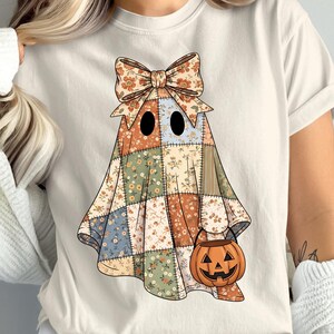 May include: Cream-colored t-shirt featuring a patchwork ghost design with a floral bow and a jack-o'-lantern bucket. The ghost's body is made of various floral patterns in orange, green, and blue. The jack-o'-lantern is orange.