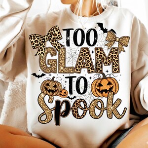 Könnte beinhalten: Ein weißes Sweatshirt mit einem Halloween-Motiv. Das Design beinhaltet den Text "Too Glam To Spook" in einem Mix aus Leopardenmuster und glitzernden goldenen Buchstaben, zusammen mit Kürbissen, Fledermäusen und Spinnweben.
