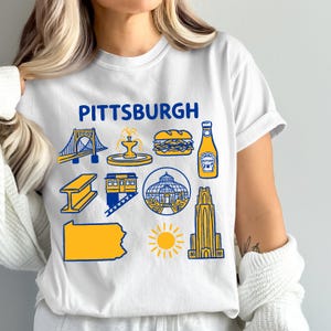 May include: White t-shirt with the word "PITTSBURGH" in blue, featuring yellow and blue illustrations of Pittsburgh landmarks, including a bridge, fountain, sandwich, ketchup bottle, and the state of Pennsylvania.