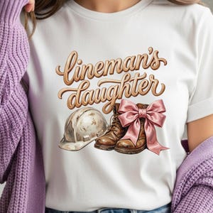 Könnte beinhalten: Weißes T-Shirt mit dem Schriftzug "Lineman's Daughter" in brauner Schrift. Das Design umfasst einen Arbeitsschutzhelm, braune Arbeitsstiefel und eine rosa Schleife.