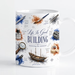 May include: A white ceramic mug featuring a colorful watercolor-style print of hobbyist tools including a magnifying glass, paintbrush, tweezers, and a model ship. The text reads Life Is Good BUILDING mastering the miniature with small phrases.