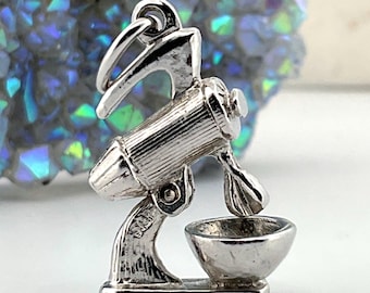 May include: A silver-toned metal charm shaped like a stand mixer, featuring a vertical motor housing, a mixing attachment, and a small bowl attached to a rectangular base. The charm includes a small circular jump ring at the top for attachment.