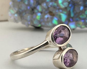May include: A silver bypass ring featuring two round, faceted purple amethyst gemstones set in polished metal bezels. The open-ended band design allows the two stones to sit side-by-side, showcasing the clear, light purple hue of the gems.