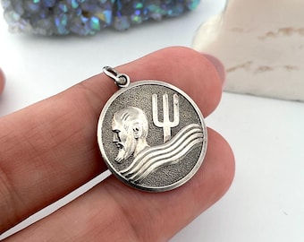 May include: A silver-toned pendant with a circular design. The pendant features a bearded man's profile and a trident symbol above wavy lines. The pendant is held by a hand against a white background, with a crystal and a white sculpture in the background.
