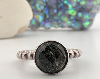 May include: A silver ring with a large, round, black gemstone. The band features a beaded design. The gemstone has a unique, textured appearance. The ring is set against a white background.