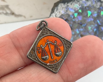 May include: Vintage silver-toned pendant with an orange enamel center featuring a Libra scale design. The pendant has a square shape with a decorative border and the word "SEPT" is visible. The pendant is held in a hand.