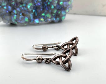 May include: A pair of silver-toned Celtic knot earrings. Each earring features a stylized triquetra knot pendant suspended from a curved ear wire with a small bead accent. The earrings are displayed on a white surface.