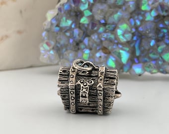 May include: A small, detailed silver-toned chest pendant with a hinged lid and decorative metalwork. The chest has a rustic, wooden appearance with metal bands and rivets. The background features a white stone and a geode with iridescent crystals.