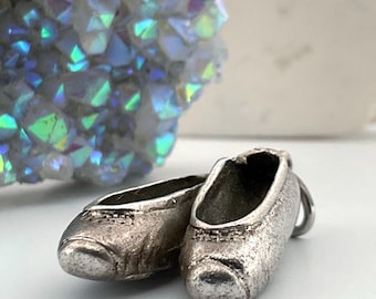 May include: A silver-toned metal charm shaped like a pair of ballet slippers. The charm features a textured, matte finish with visible stitching details along the toe and opening of the shoes. A small metal jump ring is attached to the back heel.