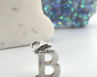 May include: A silver-colored charm in the shape of the letter "B" with a lobster clasp. The charm is displayed against a white background, with a white stone and a blue and green crystal formation in the background.