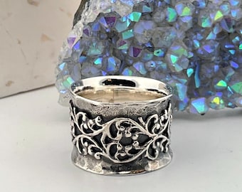 May include: A silver ring with an ornate, openwork design. The ring features a wide band with a hammered texture and a detailed floral or vine-like pattern around the center. The ring is set against a backdrop of a white stone and a geode.