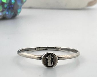 May include: A silver ring with a small, circular charm featuring the letter "t". The ring has a thin band and a simple, minimalist design. The charm is slightly darker than the band, creating a subtle contrast.
