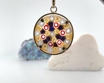 May include: A round pendant with a gold-colored frame, showcasing a colorful mosaic design. The mosaic features red, white, and blue circular patterns. The pendant is suspended from a gold-colored bail. The background is white with a few decorative stones.