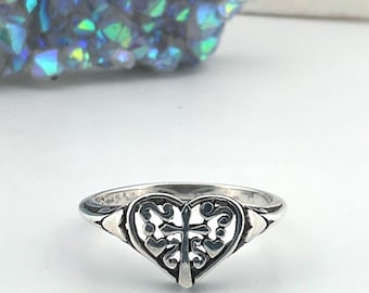 May include: A silver ring featuring a heart-shaped design with a cross in the center. The ring has intricate filigree detailing and a polished finish. The ring is set against a white background with a blurred, iridescent crystal formation in the background.