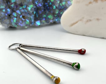 May include: Three silver-colored metal matchstick-shaped pendants with colorful tips: red, green, and yellow. The pendants are attached to a small silver ring. The background includes a geode and a white heart-shaped stone.