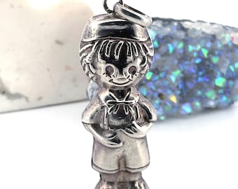 May include: A silver-toned charm in the shape of a small child, wearing a hat and shorts, holding something in front. The charm has a loop at the top for attaching to a chain or bracelet. The charm is detailed with facial features and clothing.