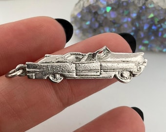 May include: A silver-colored convertible car charm with detailed features, including wheels, doors, and a convertible top. The charm is held between fingers, showcasing its size and design. The charm has a loop for attaching to a necklace or bracelet.