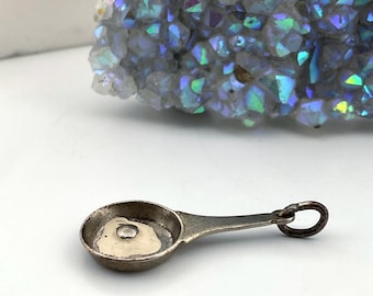 May include: A small, antique silver measuring spoon with a round bowl and a handle ending in a loop. The spoon is set against a white background, with a large, iridescent crystal formation in the background.