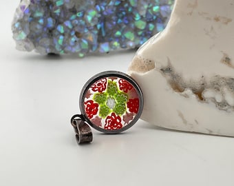 May include: A round pendant with a silver-colored metal frame. The pendant features a colorful floral design with red and green elements. The background is white. The pendant is displayed on a white surface.