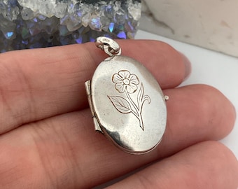 May include: Oval silver locket with a floral engraving. The locket is closed and has a small loop at the top for a chain. The flower design is etched into the front of the locket. The locket is held in a hand.