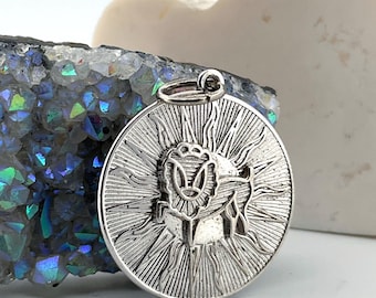 May include: A circular silver-toned pendant featuring a raised relief of a ram centered against a sunburst design with radiating rays. The pendant includes a small circular bail at the top for attachment to a chain or cord, showcasing a metallic finish.