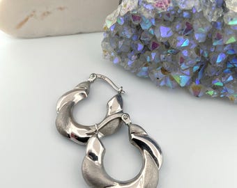 May include: A pair of silver-colored hoop earrings with a twisted design. The earrings have a matte finish on the twisted sections and a polished finish on the inner and outer curves. The earrings are displayed on a white surface.