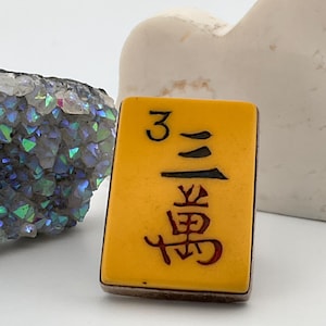 May include: A yellow Mahjong tile with black and red Chinese characters and the number 3. The tile has a dark brown border. The background is white, with a crystal and a white heart-shaped object.