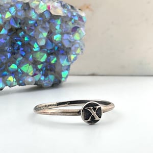 Peut inclure: Une bague en argent avec un emblème circulaire portant la lettre "X". La bague a un anneau fin et une finition légèrement oxydée. La bague est présentée sur une surface blanche, avec une formation de cristal colorée en arrière-plan.