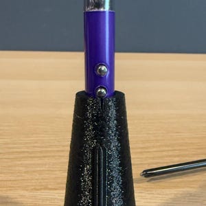 May include: A golf club grip with a black textured cone-shaped base and a purple cylindrical top. Two silver buttons are on the purple section. The grip is on a wooden surface.