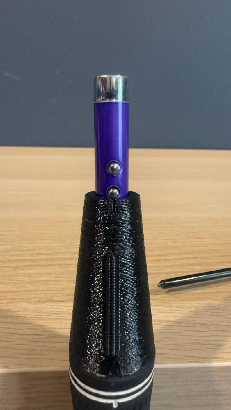 May include: A golf club grip with a black textured cone-shaped base and a purple cylindrical top. Two silver buttons are on the purple section. The grip is on a wooden surface.