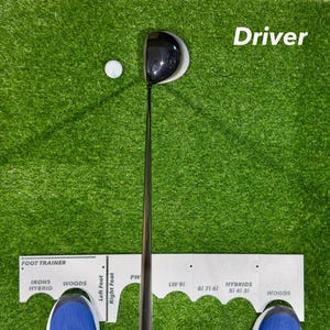 May include: A golf driver and ball are positioned on a green artificial turf. The word "Driver" is written in white. A foot trainer is at the bottom of the image, with the text "Foot Trainer" and various club types.