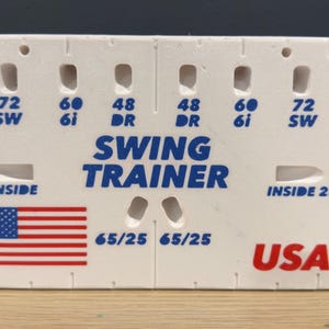 May include: White golf swing trainer with blue text reading "SWING TRAINER" and "USA". Includes the American flag and various numbers. A golf ball and tees are also visible.
